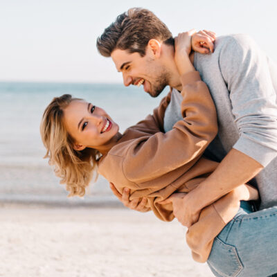 short-haired-blonde-lady-embracing-husband-beach-outdoor-portrait-good-humoured-man-dancing-with-girlfriend-near-ocean